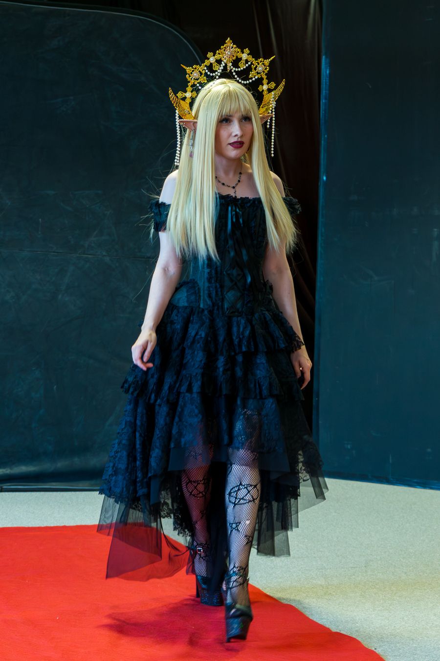 Figure in black tulle dress and crown walks across a red carpet against dark backdrop in fashion show setting.