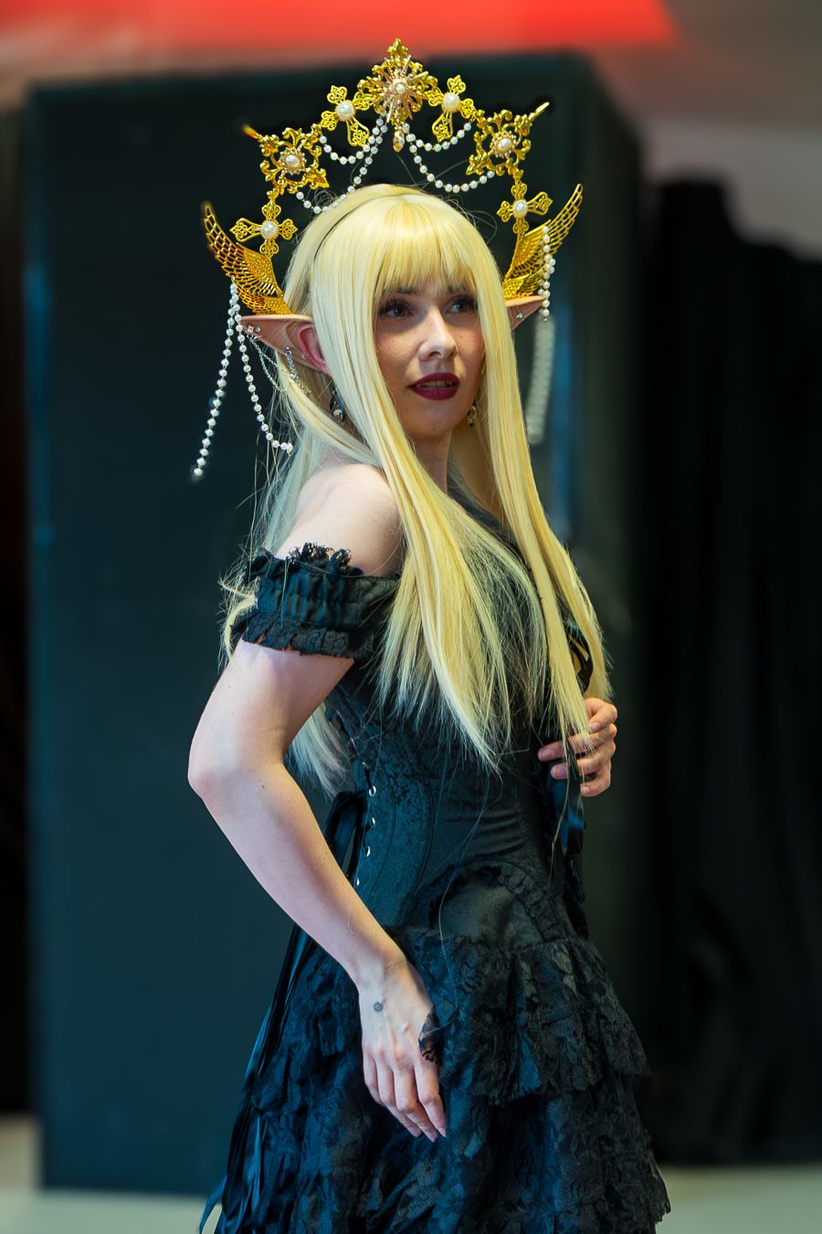 Cosplayer wearing black gothic dress and ornate gold crown with dangling jewels poses at convention event.