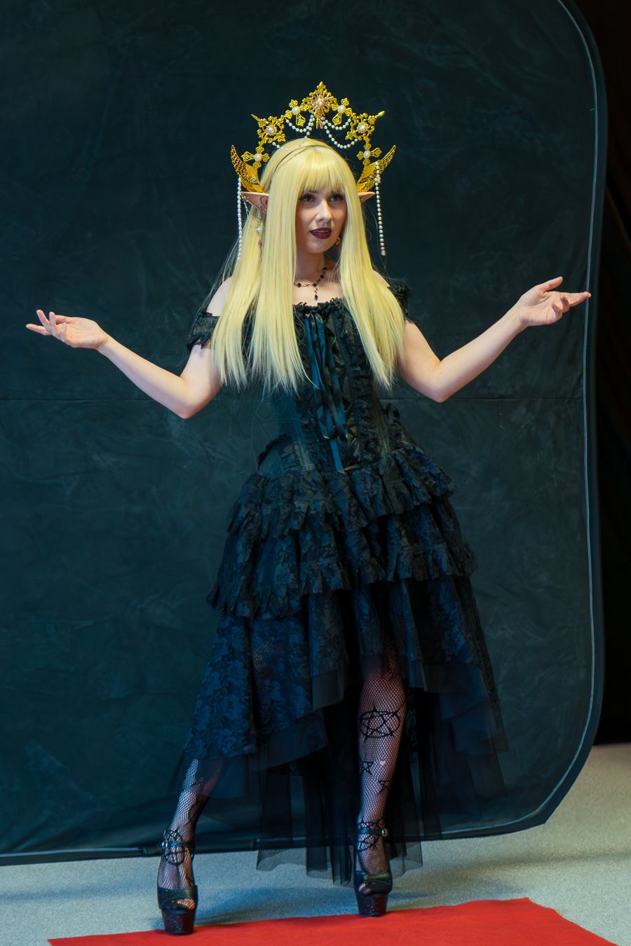 A cosplayer in a gothic black tiered dress and ornate crown poses with outstretched arms on a red carpet.