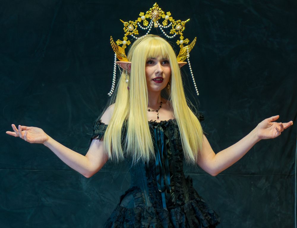 A figure in a black dress and gold crown poses with outstretched arms against a dark background.