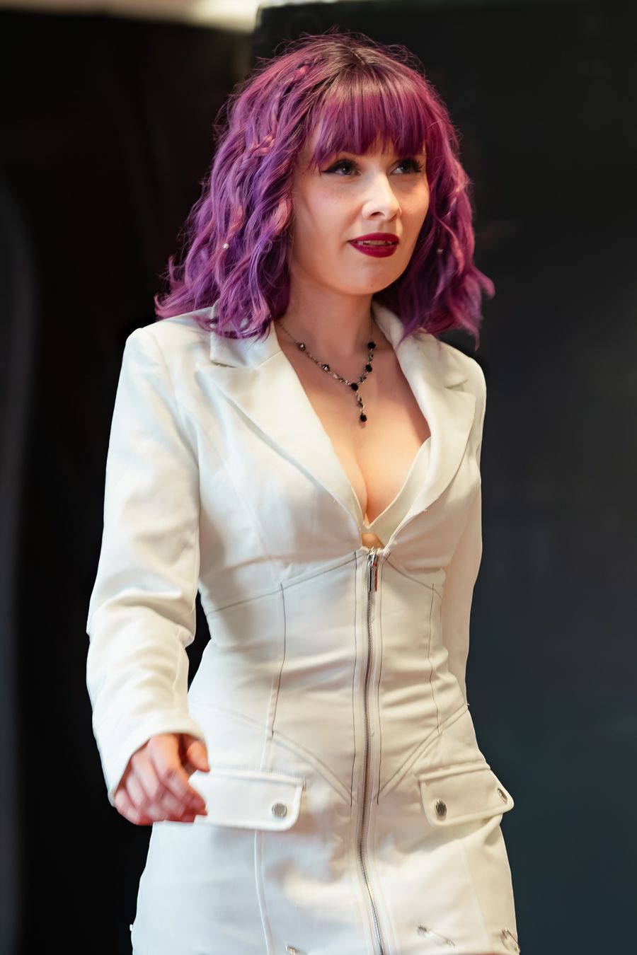 A person with vibrant purple hair wearing a white fitted jacket poses against a dark background.