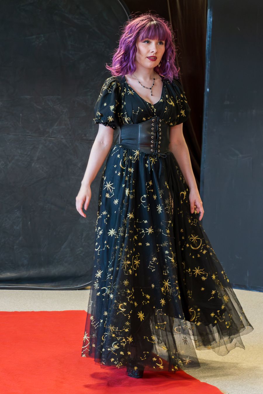 A person wearing a black celestial-patterned gown with gold stars and a corset waist stands on a red carpet.