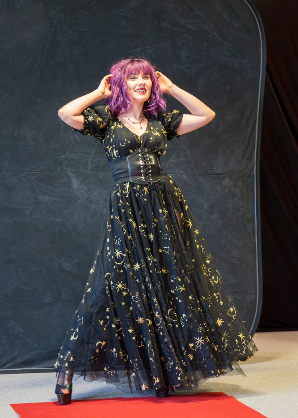 A performer in a black sparkly dress with pink hair poses dramatically on a red carpet during a theatrical performance.