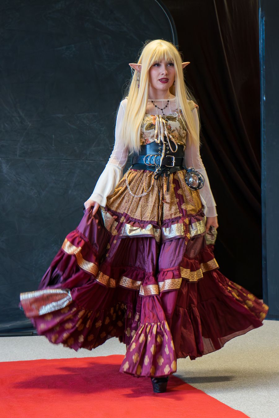 Cosplayer in an elaborate burgundy and gold steampunk-inspired dress twirls on a red carpet at a convention.