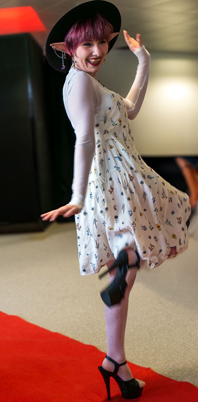 A person in a white patterned dress and black hat twirls playfully on a red carpet, showing off their purple hair and platform heels.