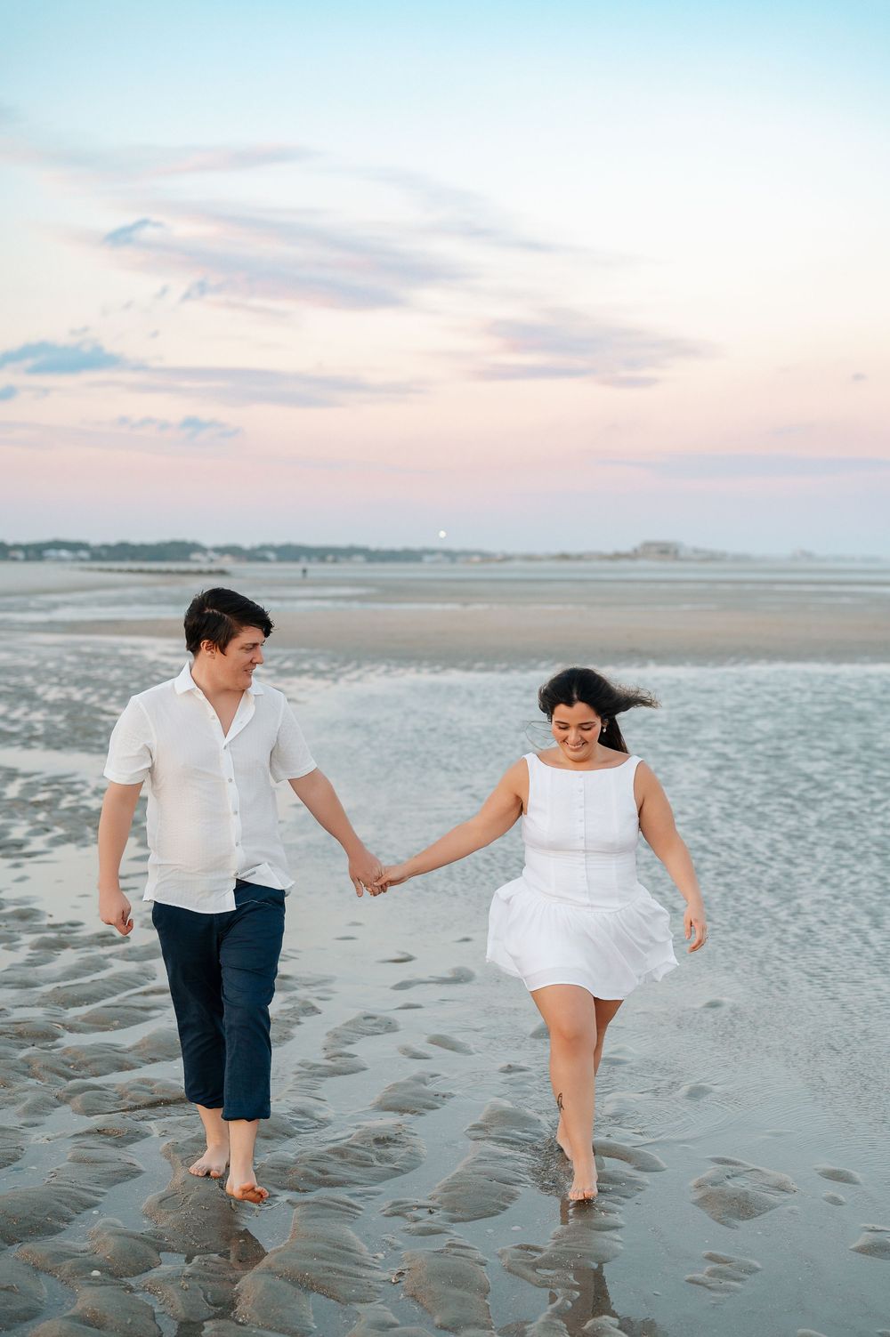 Couple holding hands while walking barefoot on beach during sunset in white outfits.
