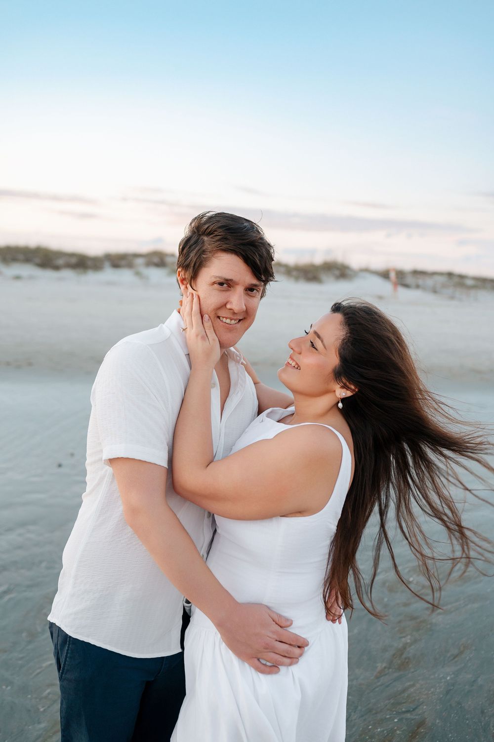 Couple in white clothing sharing playful moment on beach with wind-blown hair.