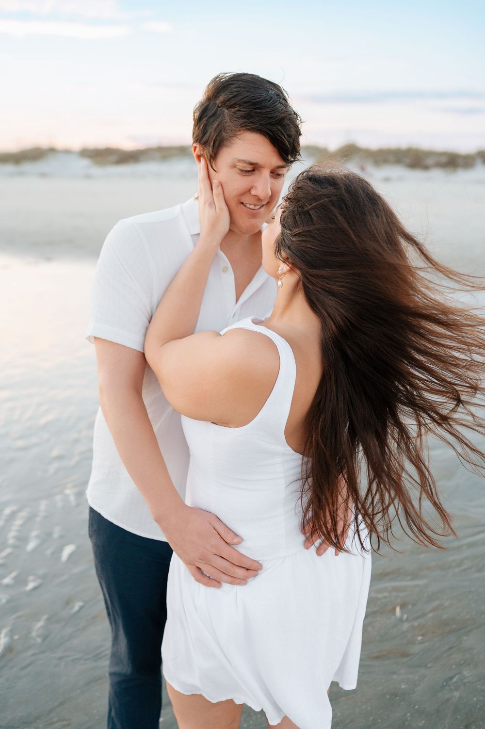 Intimate beach portrait of couple in white clothing with wind-swept hair at sunset.