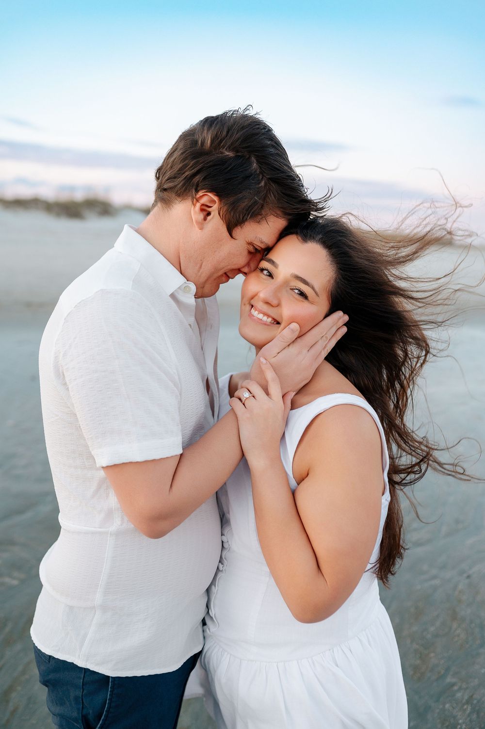 Romantic couple shares intimate moments on a beach at sunset wearing white and blue casual outfits.