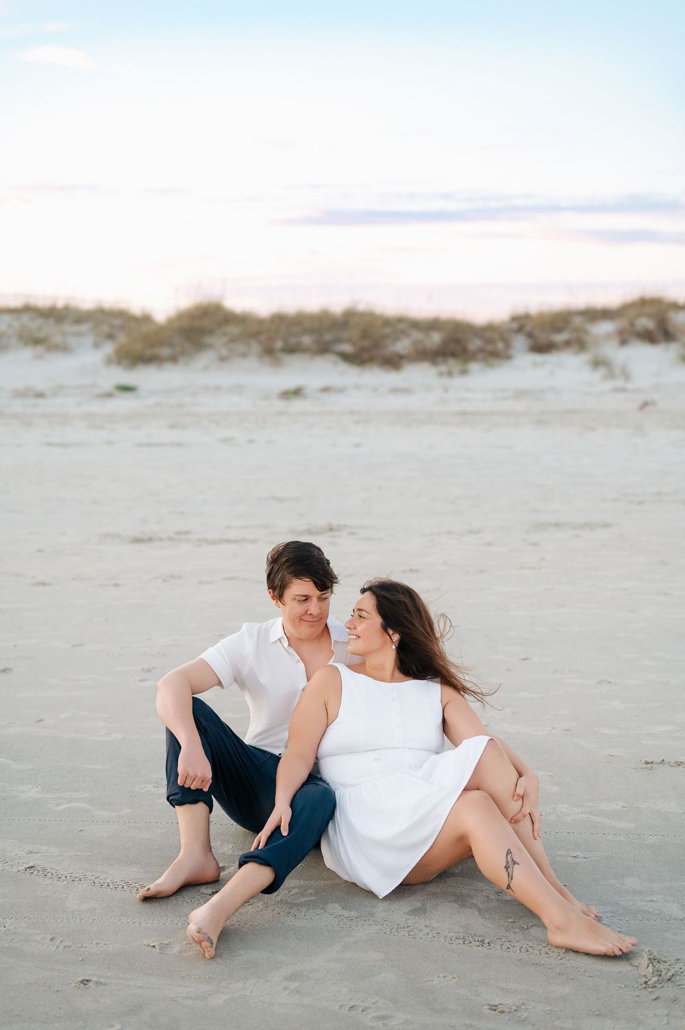 Couple relaxes together on sandy beach during casual photoshoot in matching white outfits.