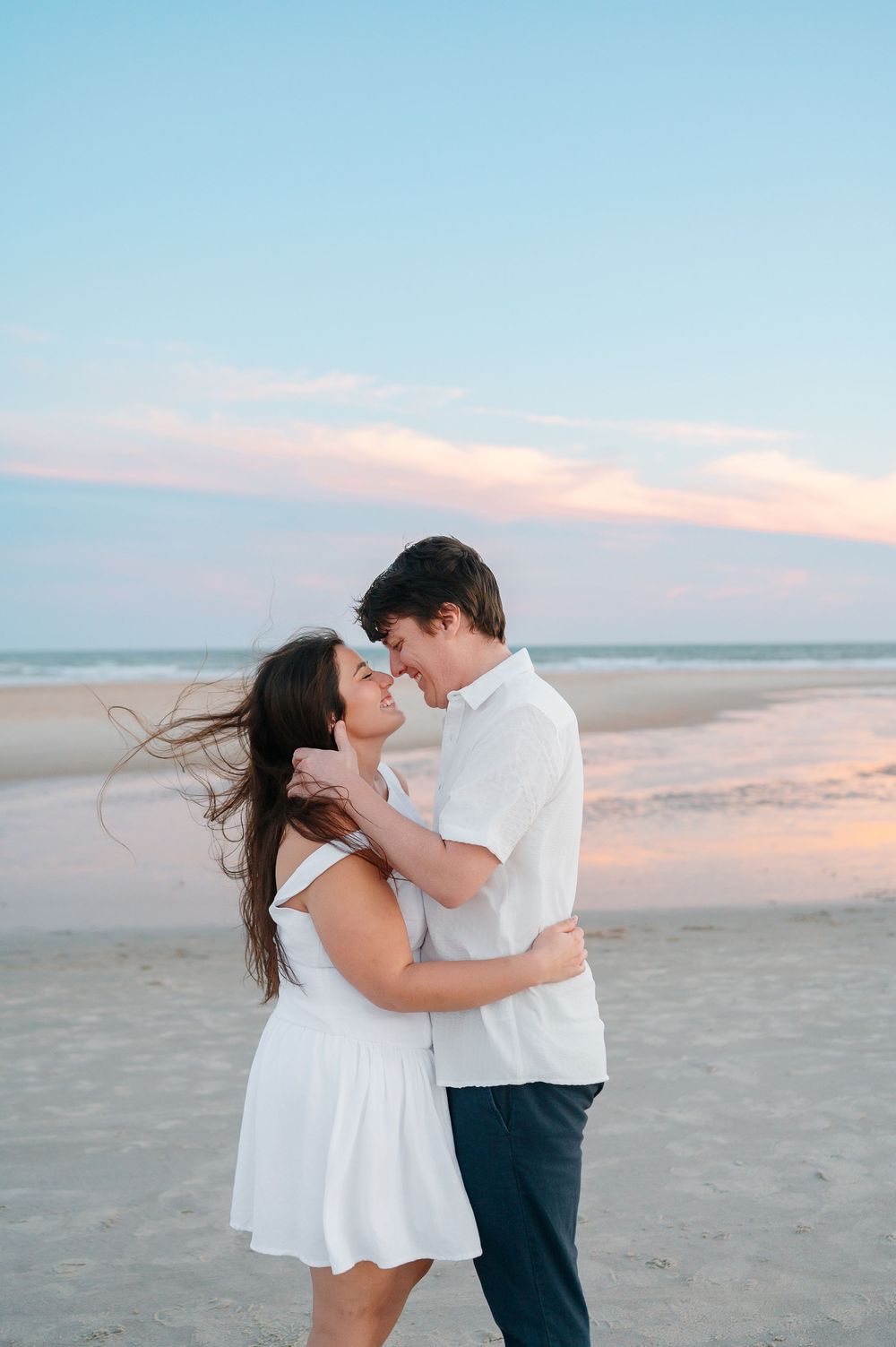 Couple embraces on empty beach during sunset wearing coordinated white and navy outfits.