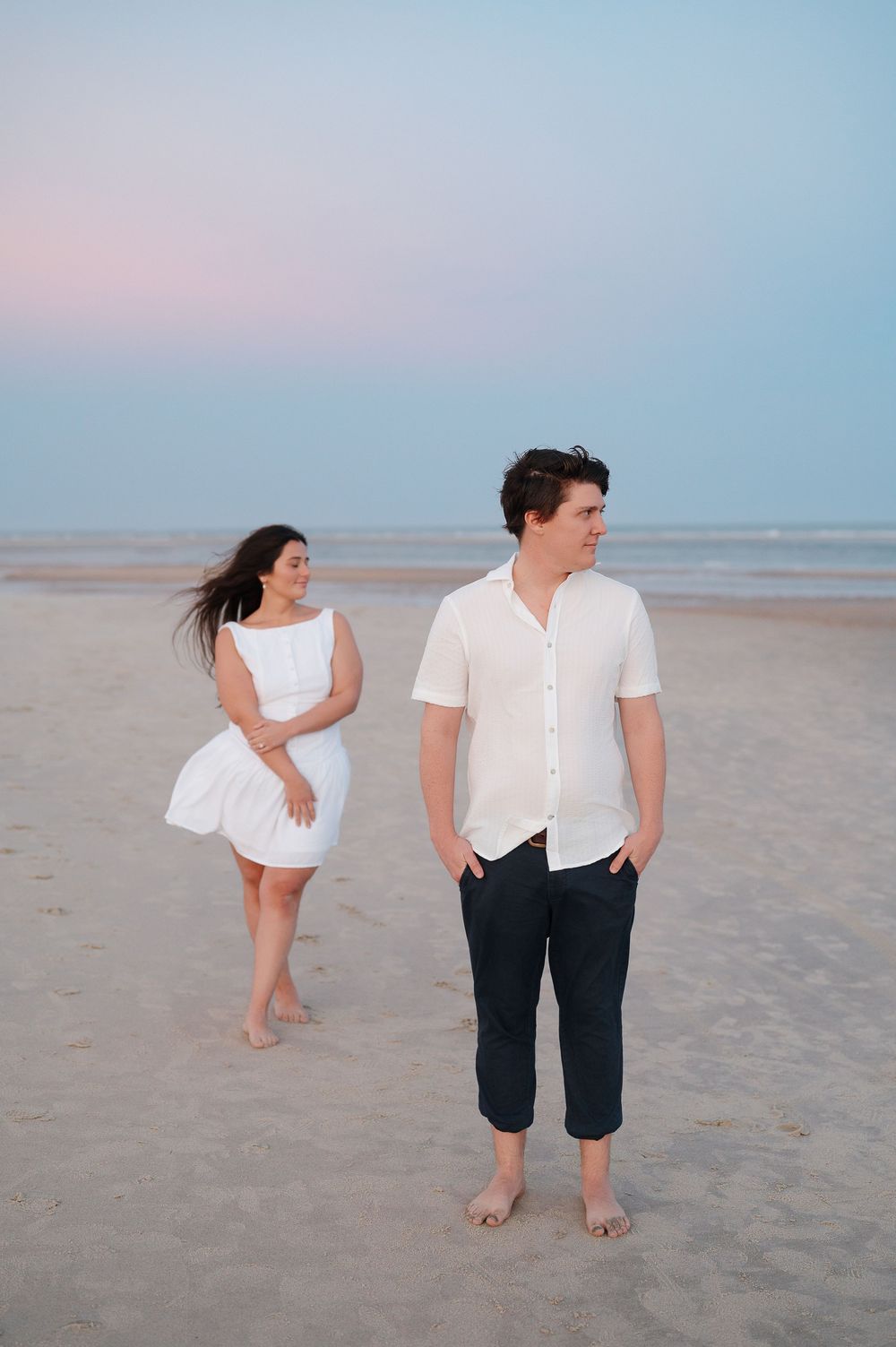 Playful couple dances and poses on sandy beach wearing casual white and navy outfits.