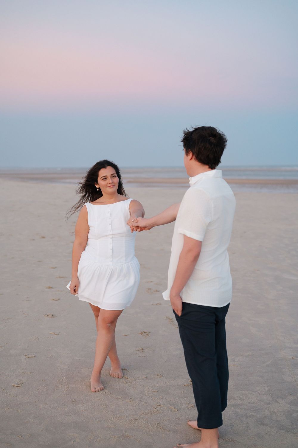 Couple shares playful dance moves on beach at dusk in coordinated white and navy attire.