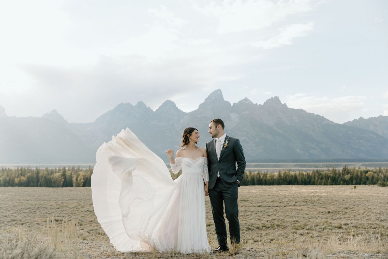 A couple in wedding attire stands in a scenic mountain meadow as the bride's dress flows in the wind.