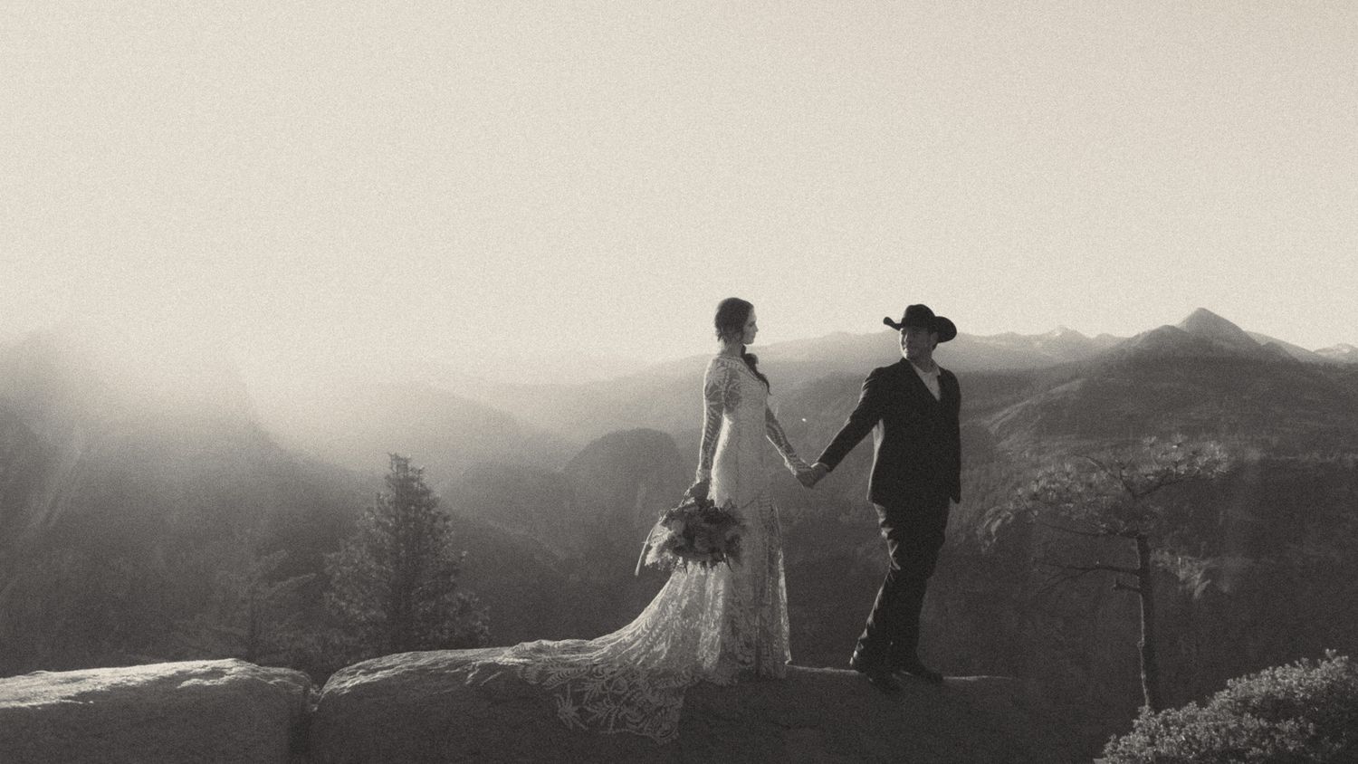 A couple in wedding attire stands silhouetted against a misty mountain landscape at sunset.
