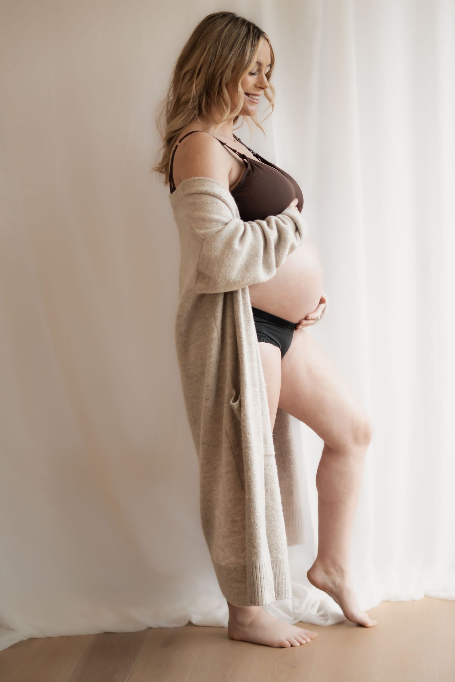 Artistic maternity photos showing pregnant woman in beige cardigan against white backdrop. Photography in Buckinghamshire.