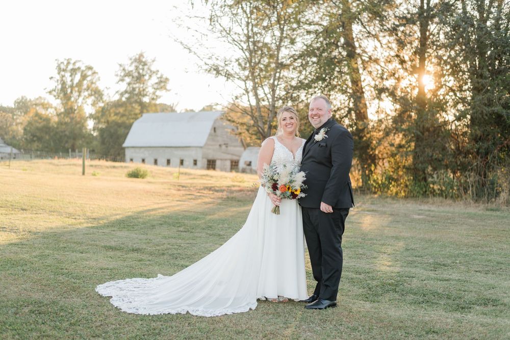 Bride and groom pose at golden hour near a rustic barn, she holding a colorful bouquet in a flowing white gown.