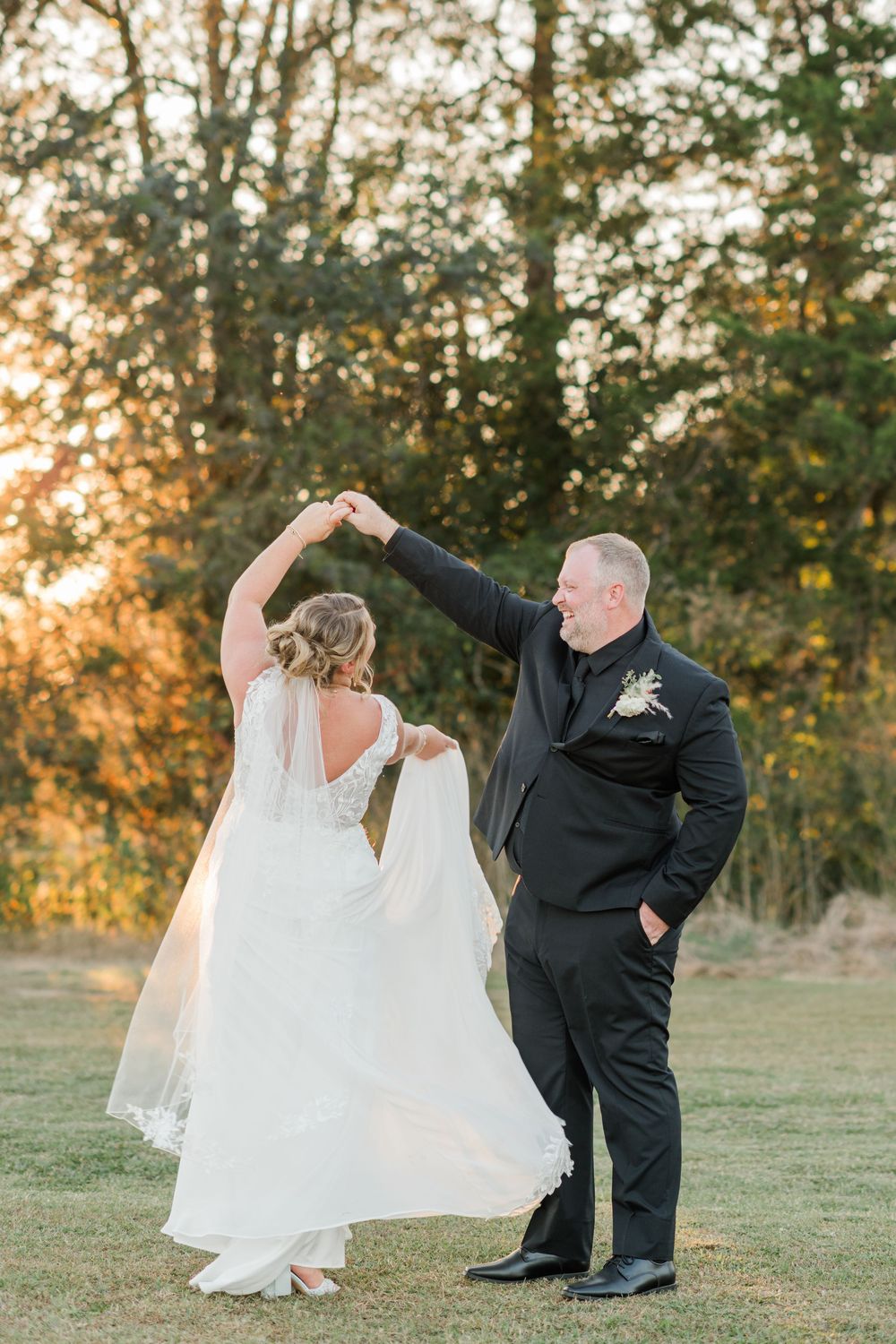 Groom twirls bride in white lace gown at sunset, both smiling joyfully on a green lawn with trees behind.
