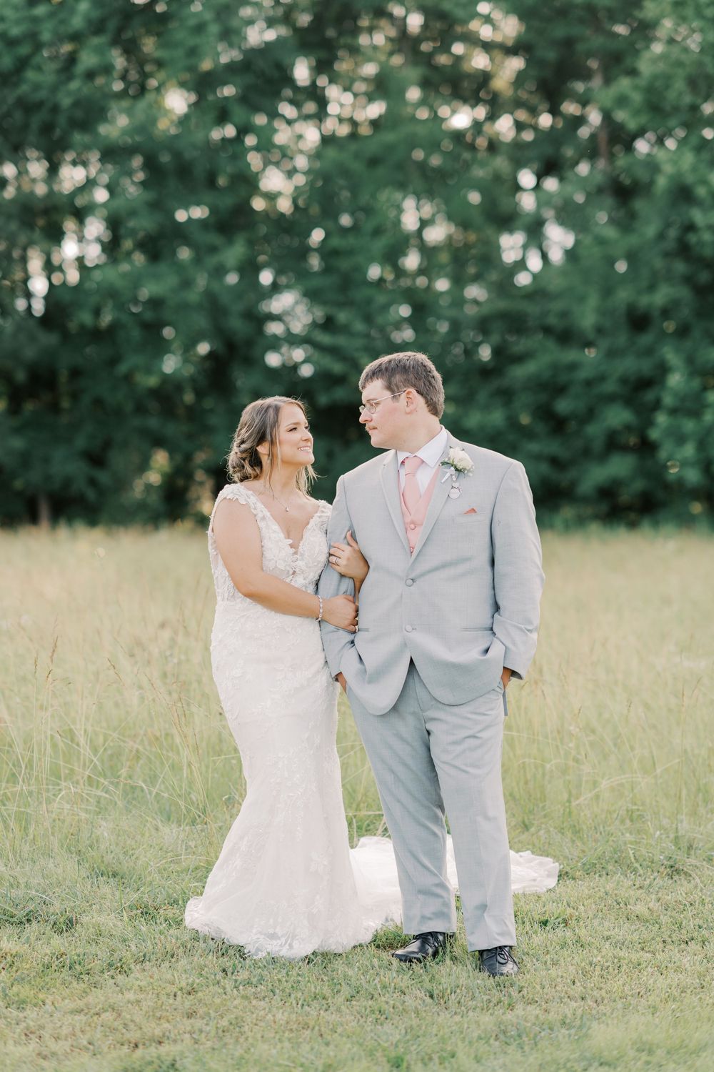 Bride and groom share a tender moment in a lush green field, she in lace gown, he in gray suit with pink tie.