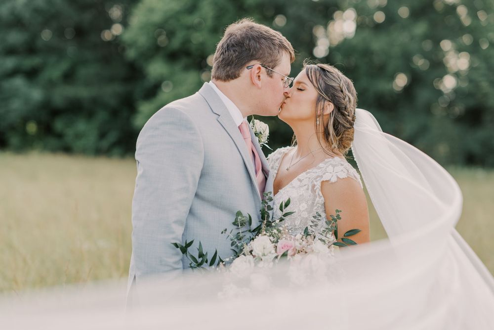 Bride and groom share a romantic kiss outdoors, her veil flowing in the breeze as she holds a pink bouquet.