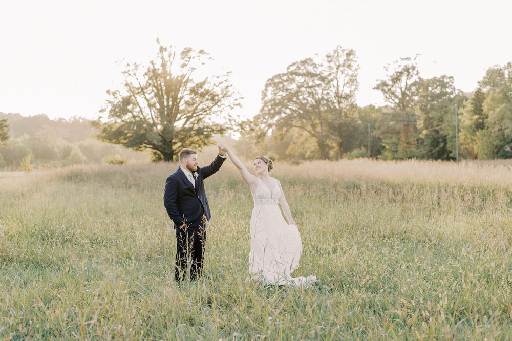Groom twirling bride in a sweeping lace gown in a sun-drenched grassy field at sunset.