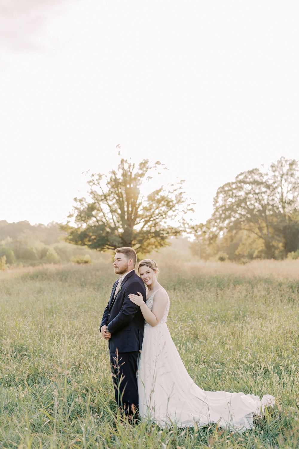 Bride resting her head on groom's shoulder in a glowing golden meadow during wedding portraits.