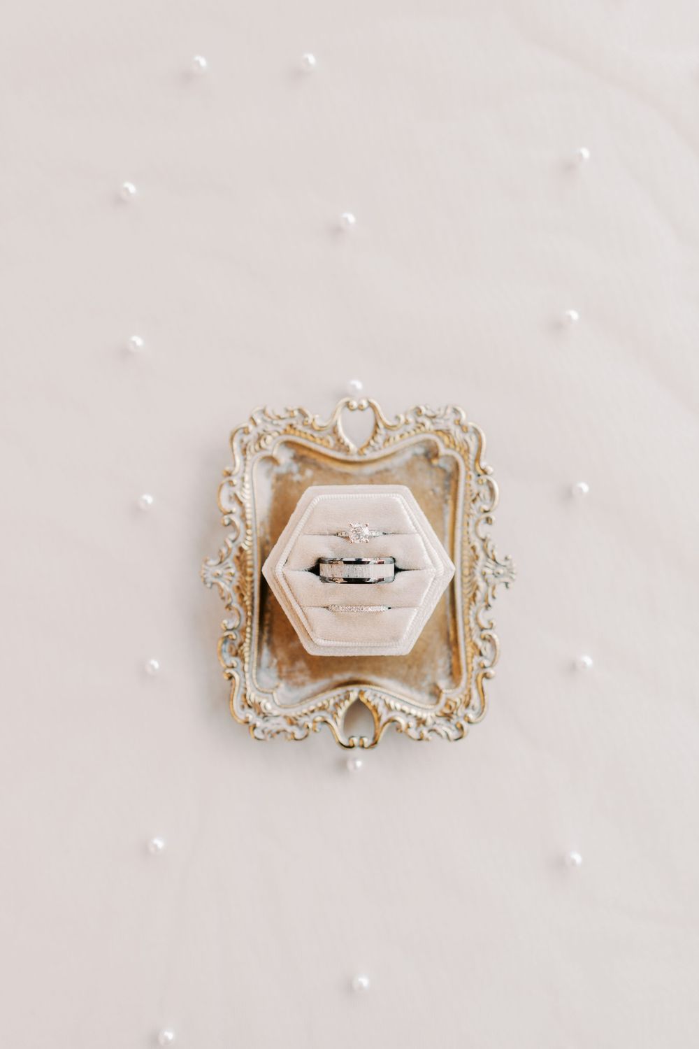 Wedding rings displayed in hexagonal velvet box on ornate gold tray surrounded by scattered pearls.