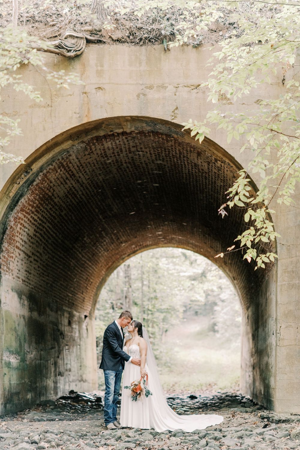 Bride and groom sharing romantic moment under historic brick tunnel arch surrounded by autumn foliage.