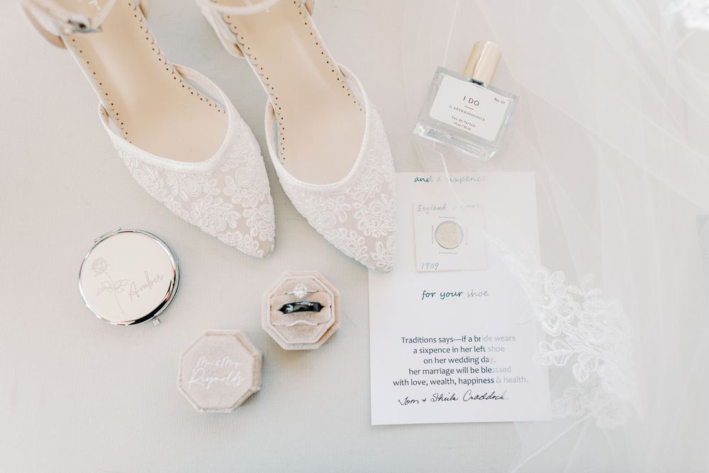 Bridal flat lay with lace heels, ring boxes, perfume bottle, and wedding invitation on white surface.