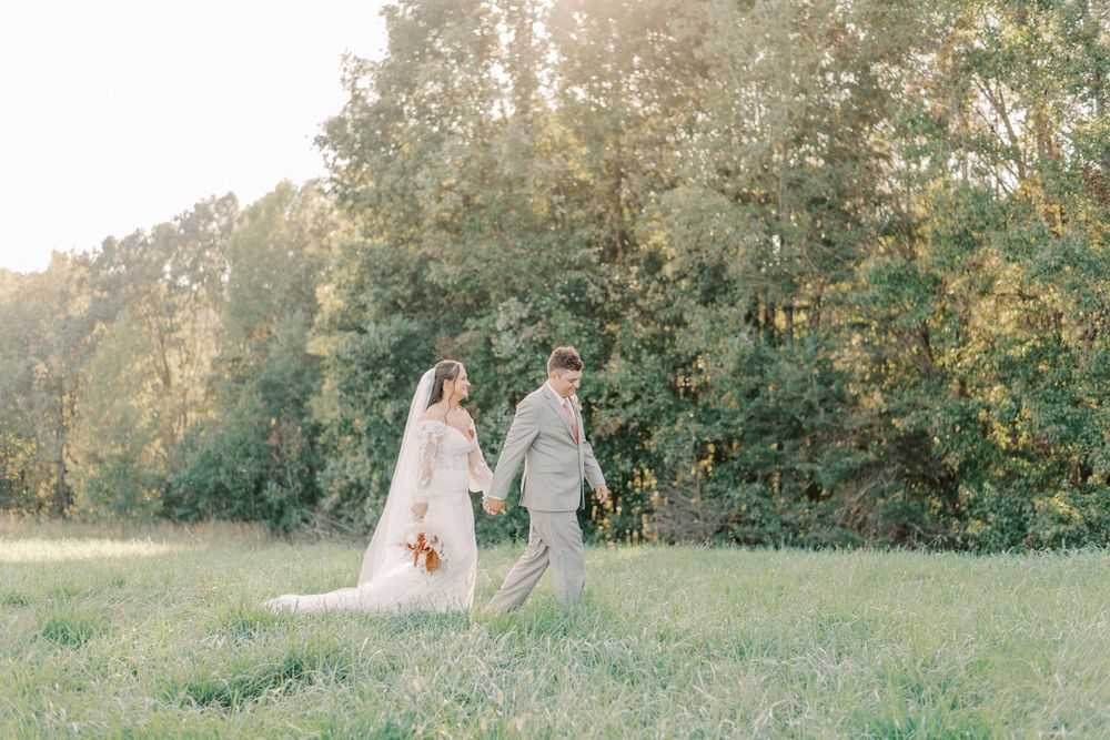 Bride and groom walking hand in hand through a sunlit green meadow on their wedding day.