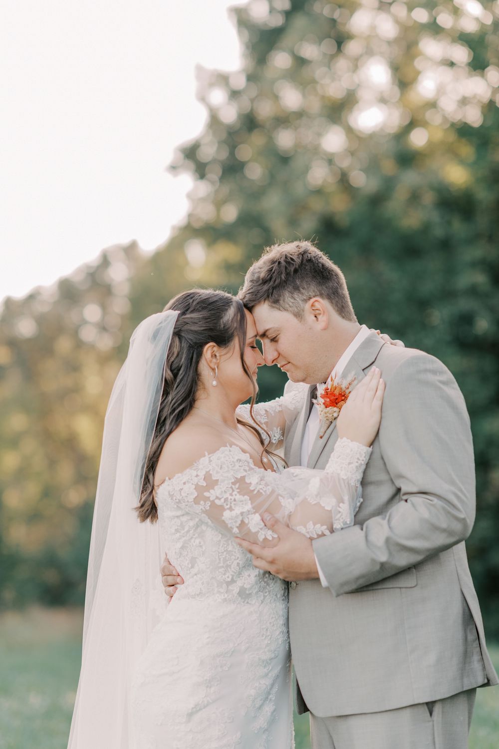 Bride and groom touching foreheads tenderly in a sunlit outdoor setting on their wedding day.