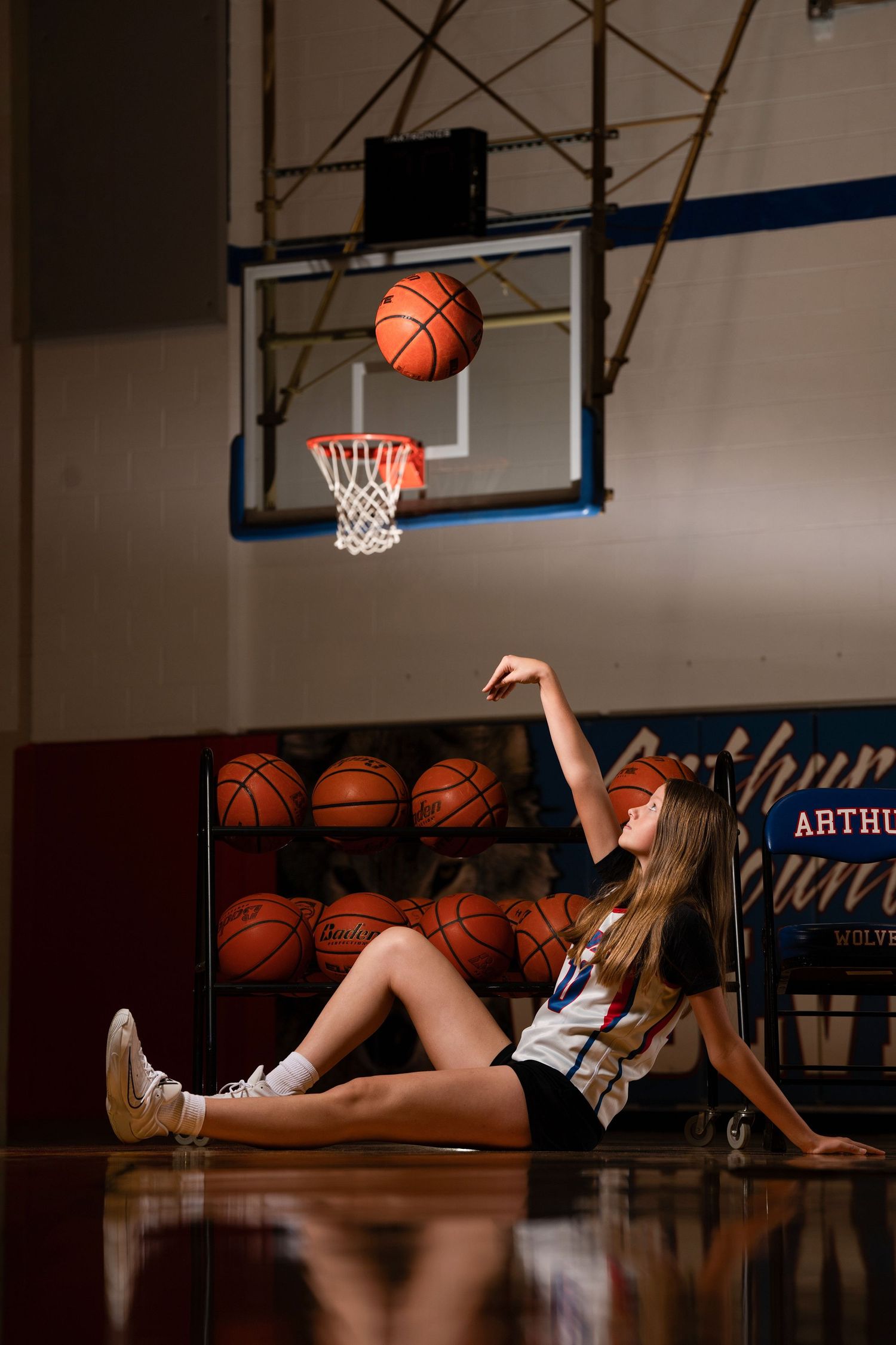 Basketball player relaxes on gym floor with rack of basketballs while ball hovers above basketball hoop.