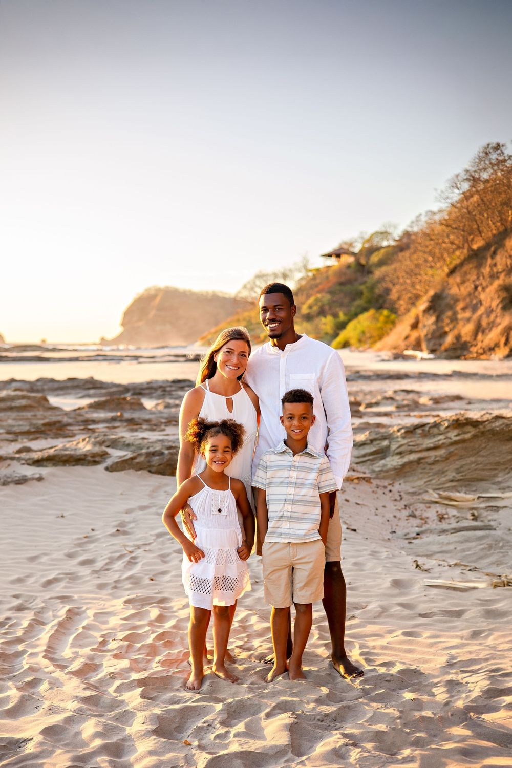 A family wearing white outfits poses together on a scenic beach at sunset with cliffs and ocean waves in the background.