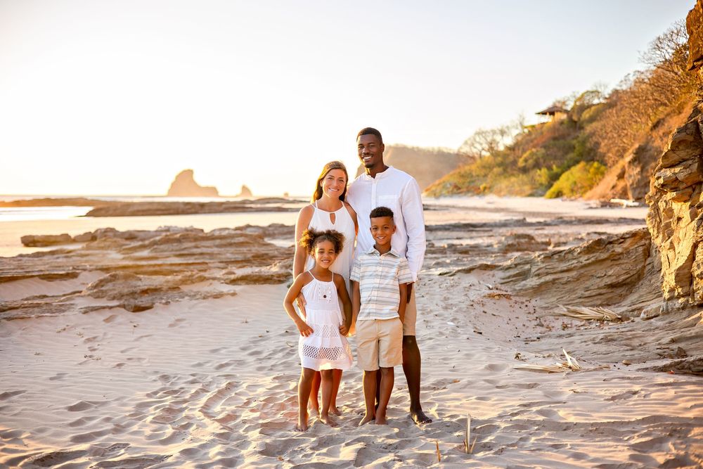 A family in white outfits poses together during sunset at a scenic beach with golden cliffs and ocean waves.