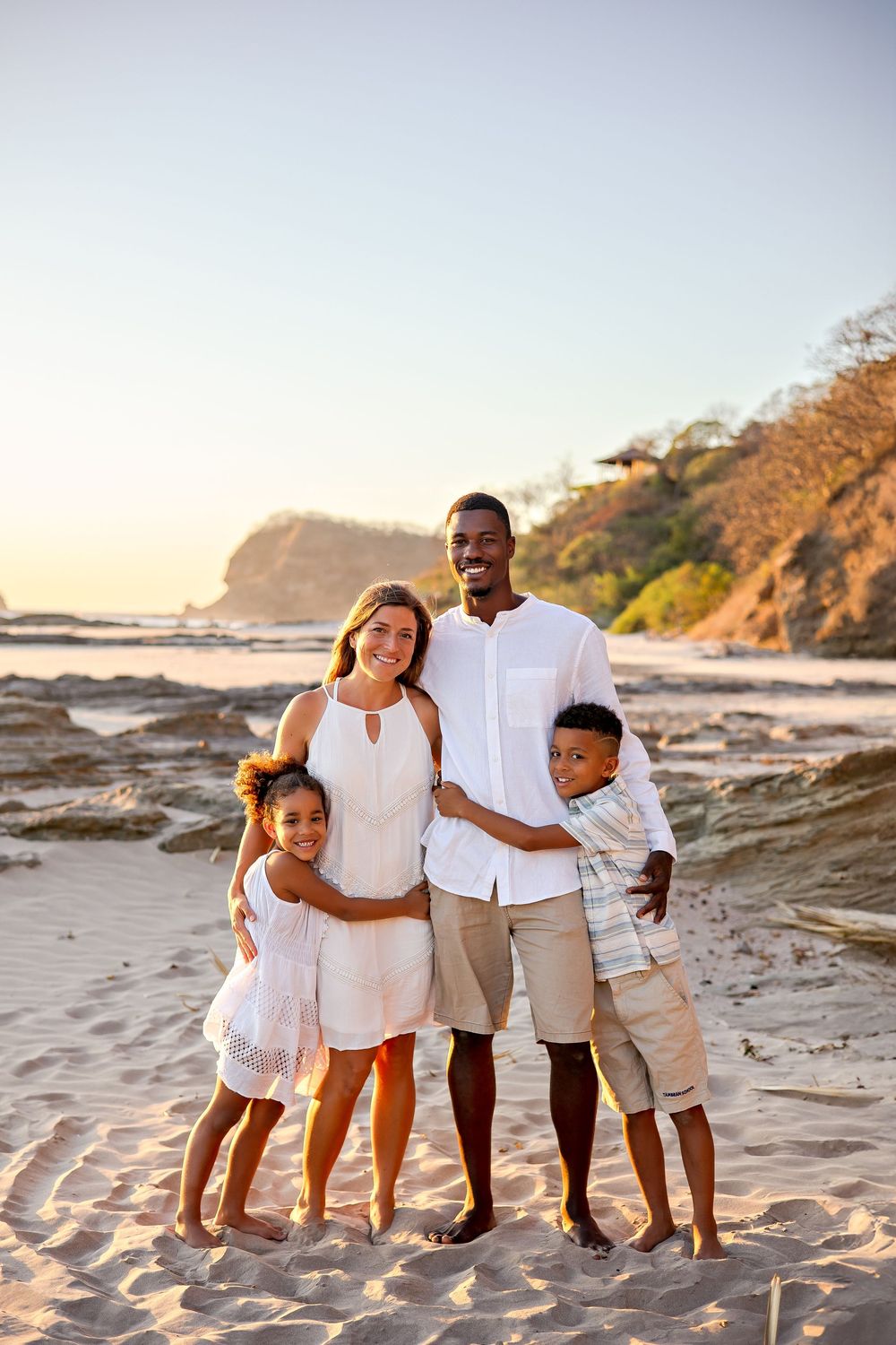 Family wearing coordinated white outfits poses together on sandy beach at sunset.