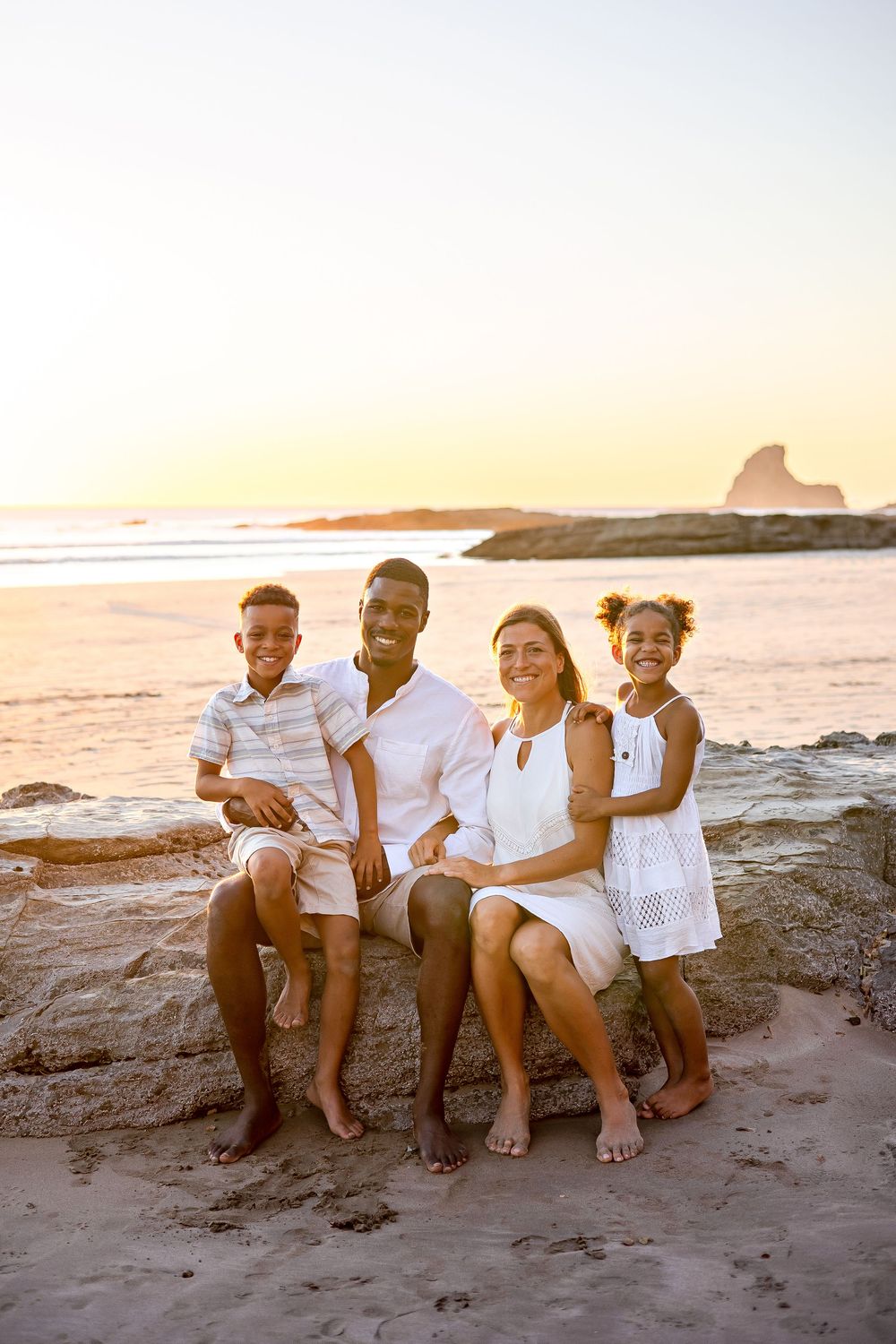 A family sits together on a beach at sunset with ocean waves and rocky coastline in the background.