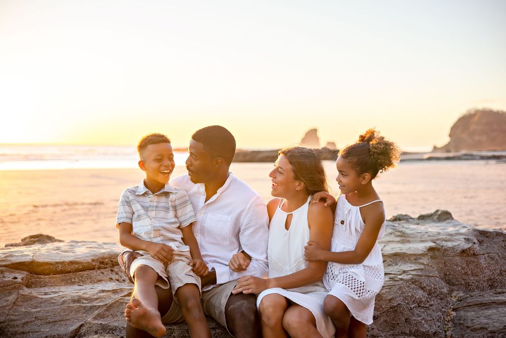 A family in white outfits sits together on rocky beach cliffs during golden hour sunset.