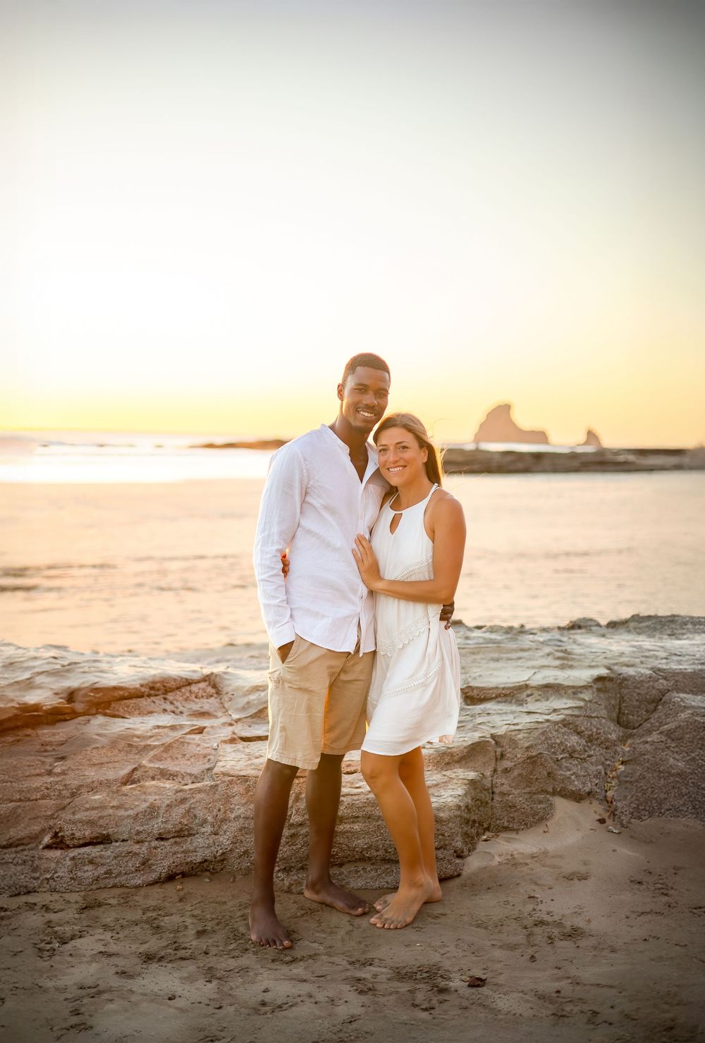 A couple in white outfits stands together on rocky beach at sunset with ocean waves and rock formations in background.