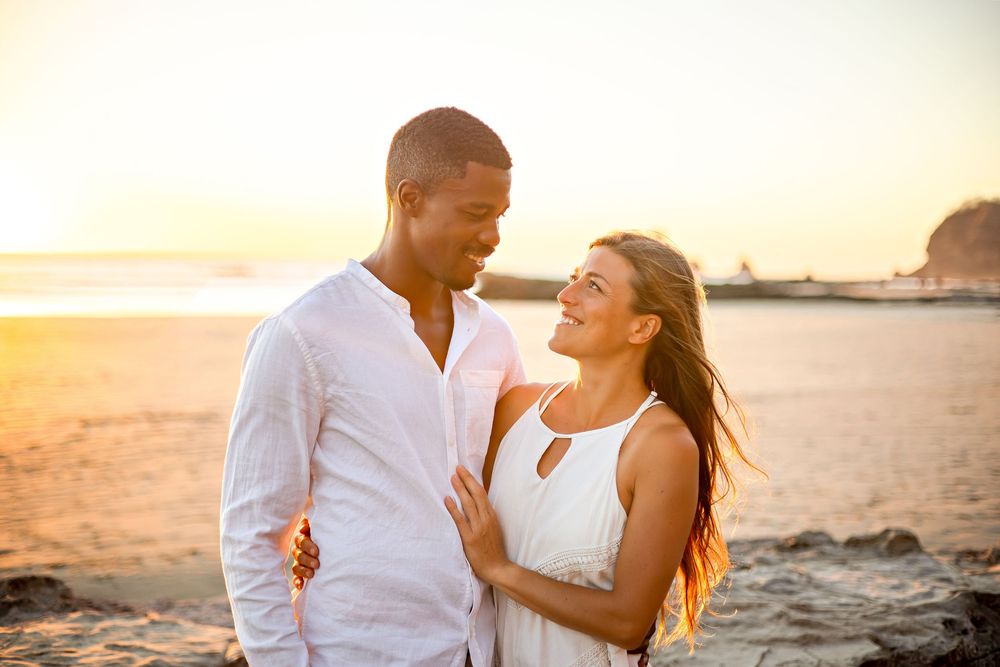 A couple in white outfits share a romantic moment on the beach at sunset.