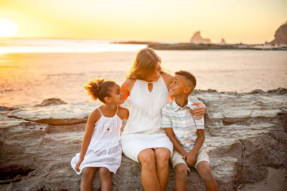 Two people in white outfits share an intimate moment on rocky beach during golden sunset.