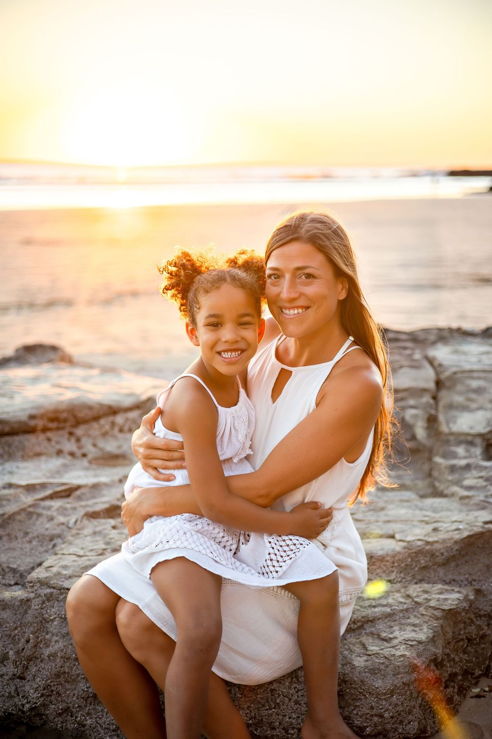 Two people in white outfits share joyful embraces while sitting on rocky coastal terrain at sunset.
