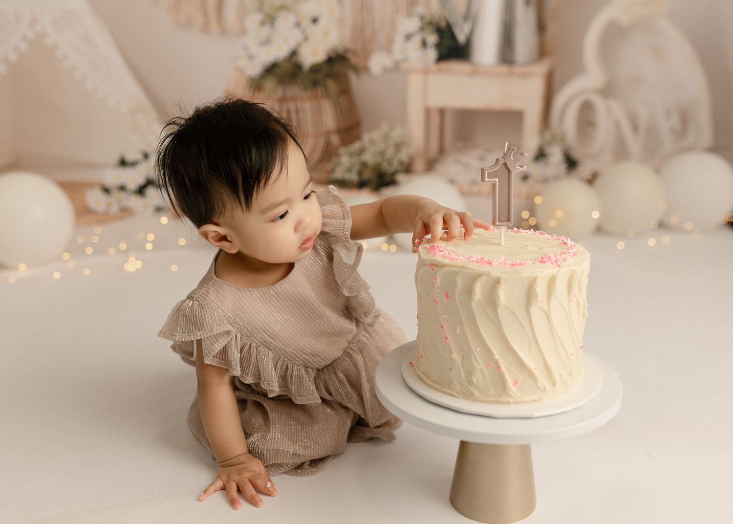 A series of photos showing a baby in neutral tones exploring and reaching for a white birthday cake on a light backdrop.