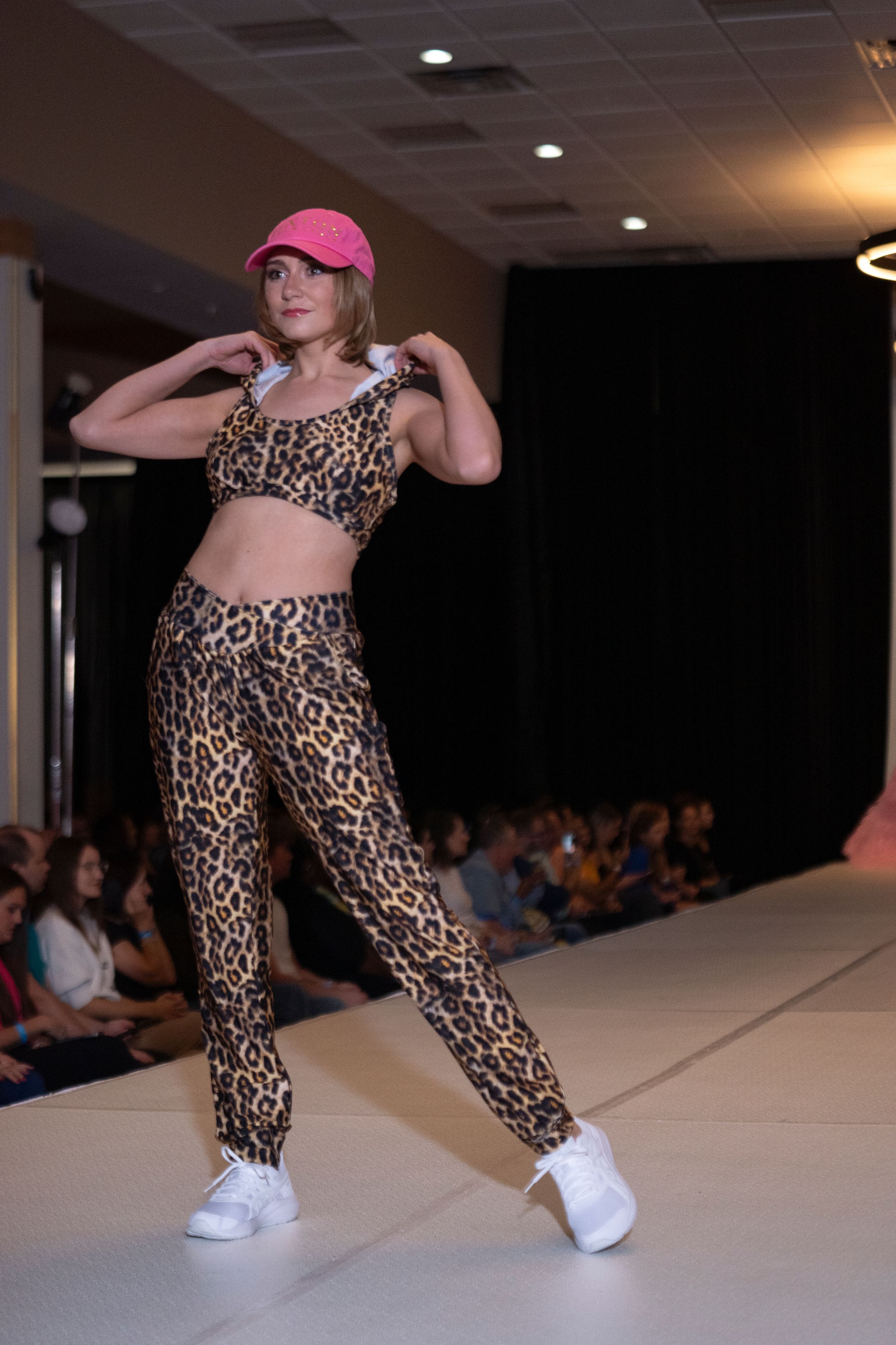 Model walks runway in leopard print jumpsuit and pink baseball cap during fashion show.