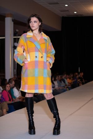 Fashion model walks runway wearing colorful plaid jacket and black boots at indoor fashion show.