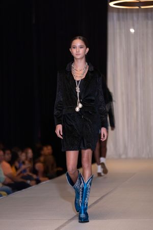 Model walks runway in black outfit paired with metallic blue knee-high boots at fashion show.