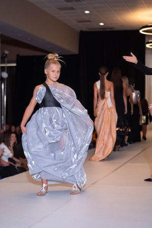 Model showcases flowing silver dress while crouching on fashion runway.