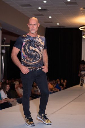 Model wearing dark dragon graphic t-shirt and jeans performs dynamic pose on runway.