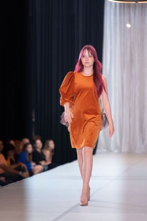 Model walks runway in vibrant orange shift dress during fashion show.