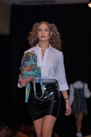 Model presents white shirt with black shorts and turquoise accessories on fashion runway.