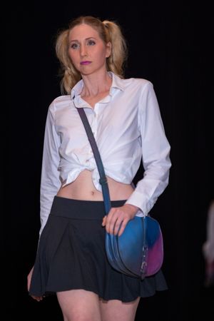 Model in schoolgirl-inspired outfit with tied white shirt and navy skirt carries blue crossbody bag.